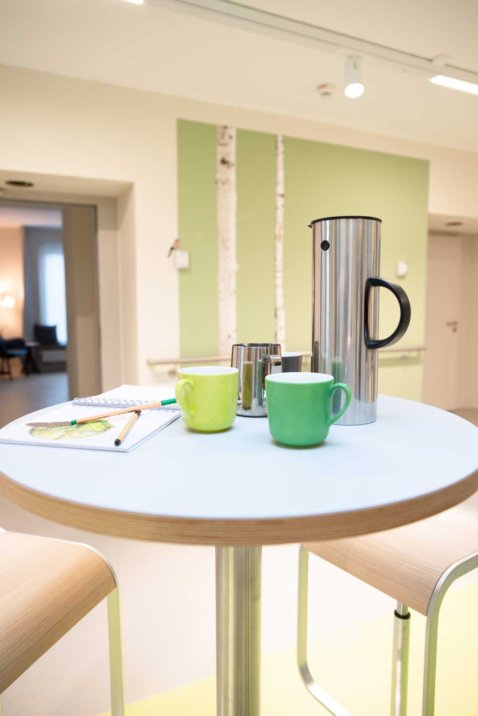 Wilkening_2OG_Tresen_Stehtisch Stehtisch im Tresenbereich mit grünen Kaffeetassen. Im Hintergrund ein Blick in ein Bewohnerzimmer